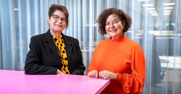 Bild på två kvinnor som står lutade mot en rosa bordskiva. De ler och tittar in i kameran. Den vänstra kvinnan bär glasögon och gul tröja med kavaj. Den högra har en orange tröja.